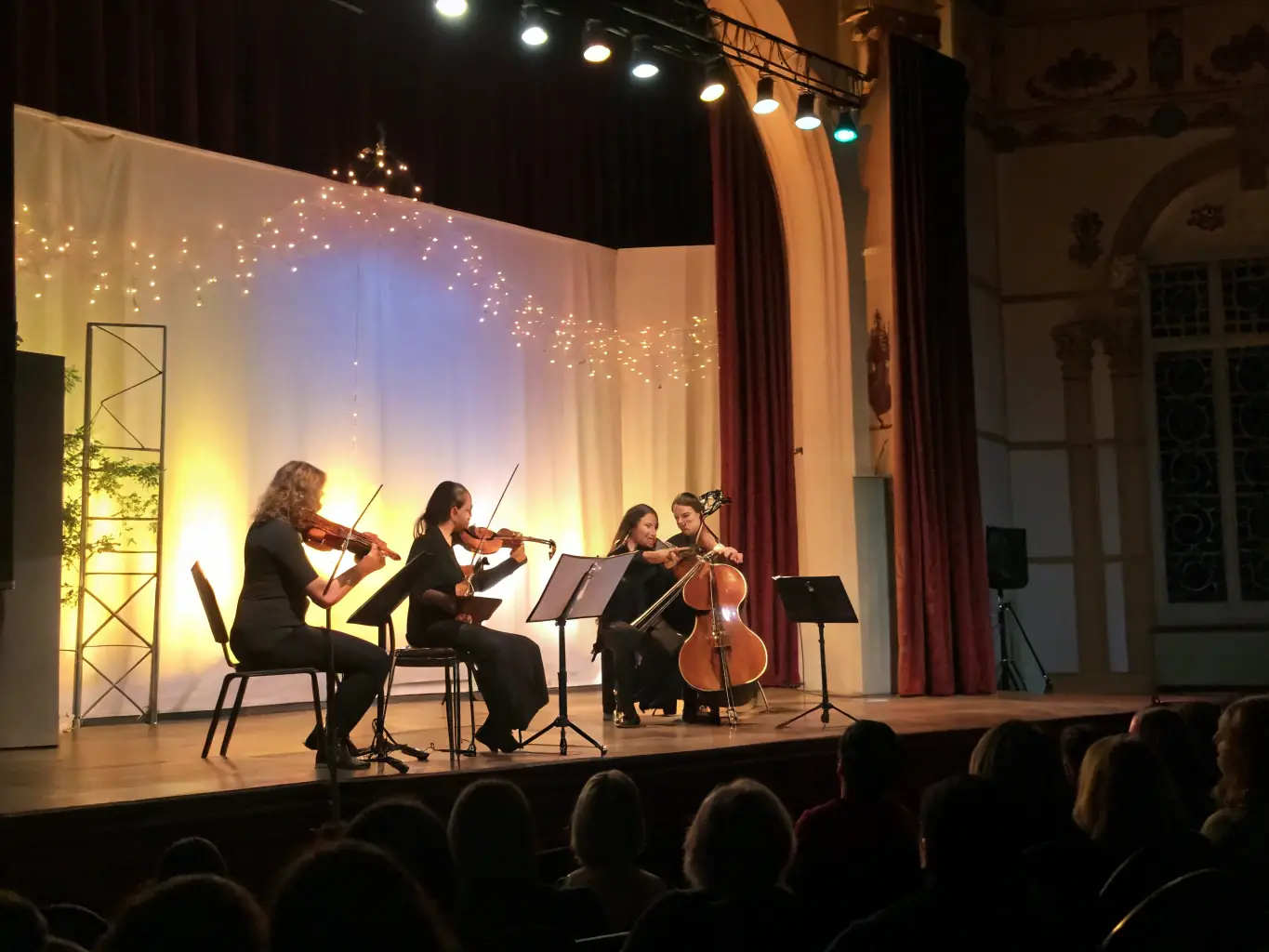 A photograph capturing a string quartet performing in an intimate setting, with audience members of all ages attentively listening. The lighting is warm and inviting, highlighting the musicians' expressions and the beauty of the music.
